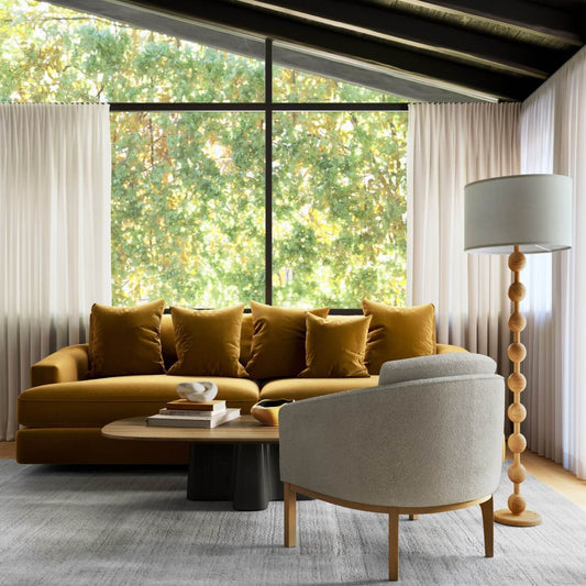 Modern living room with gold velvet sofa, gray accent chair, floor lamp and large window overlooking leafy trees