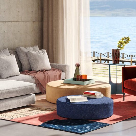 Modern coastal living room with grey sofa, round ottomans, red & blue rugs, and ocean view through window