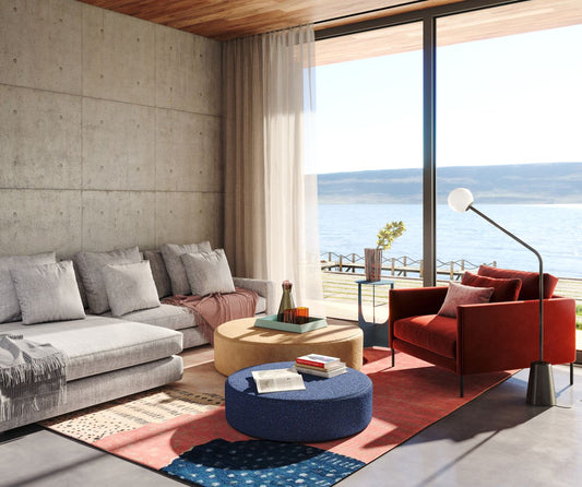 Modern coastal living room with gray sofa, red chair, ocean view, concrete walls, and colorful round ottomans