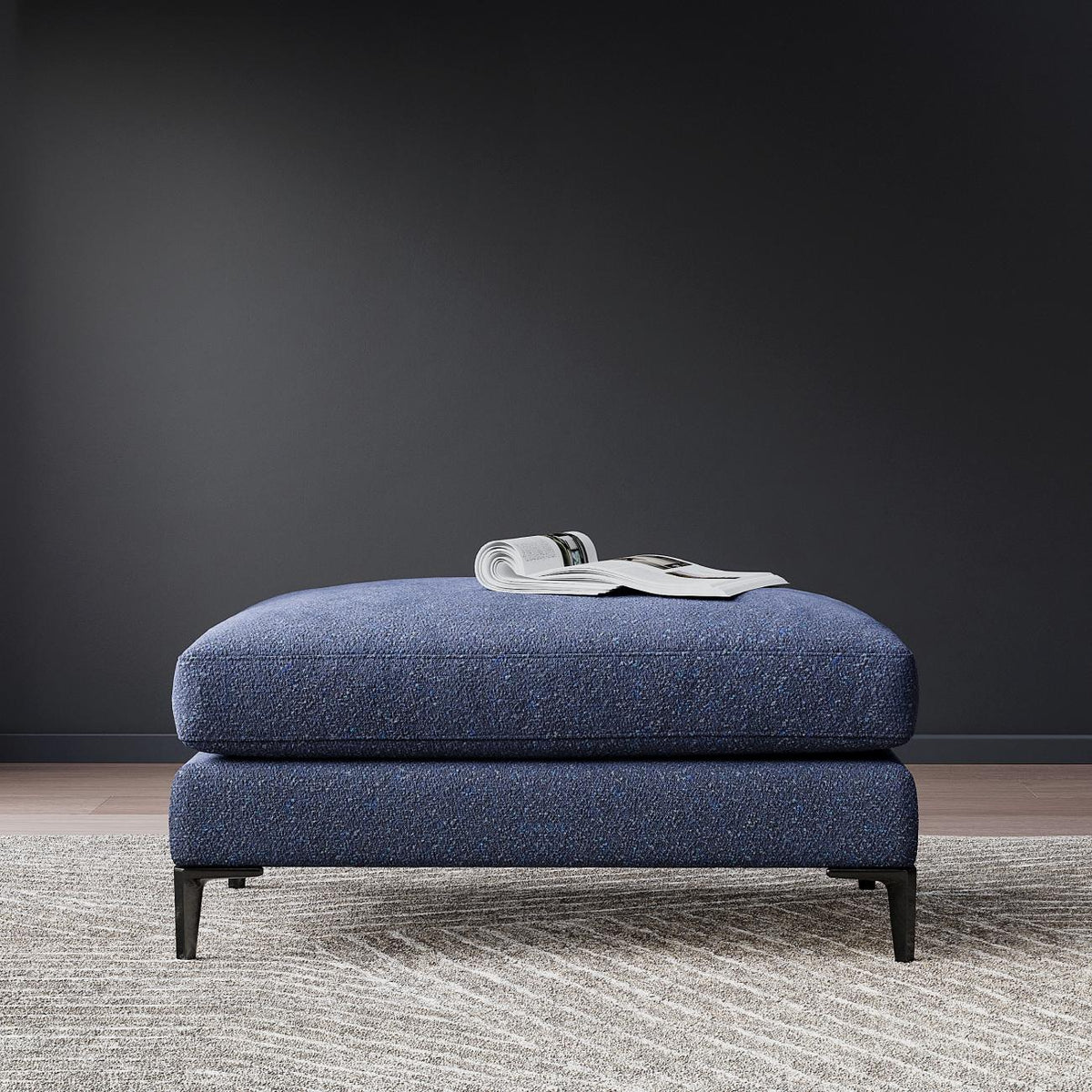 Blue upholstered ottoman with metal legs on gray carpet, open magazine on top, modern minimalist style
