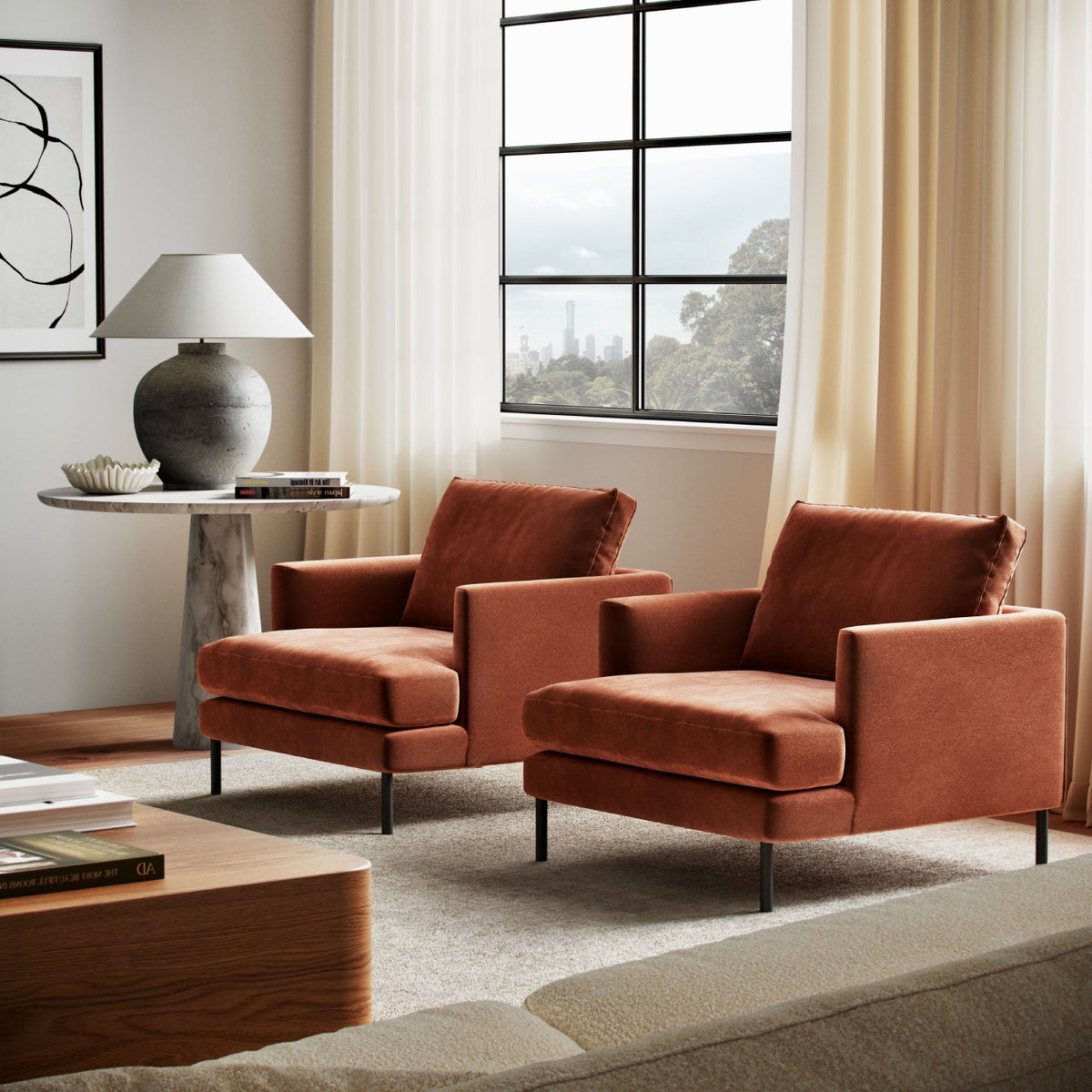 Two rust-colored velvet armchairs in modern living room with marble side table, gray lamp and city view window
