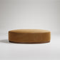Large round camel-colored velvet ottoman with dark wood base, photographed on white background
