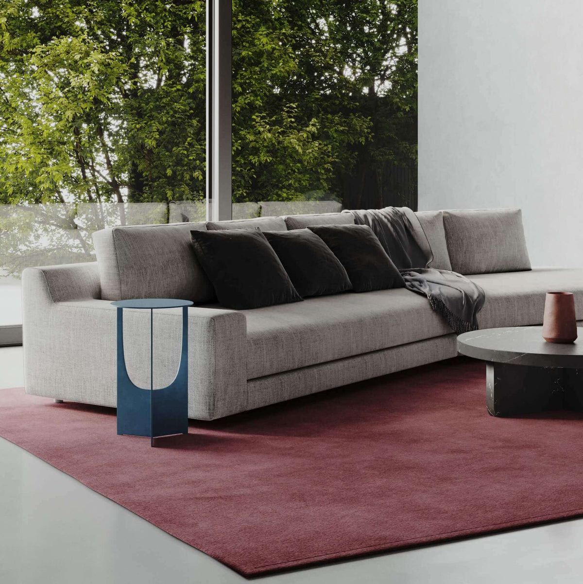 Modern gray sofa with black pillows on burgundy rug, blue side table, floor-to-ceiling windows with garden view
