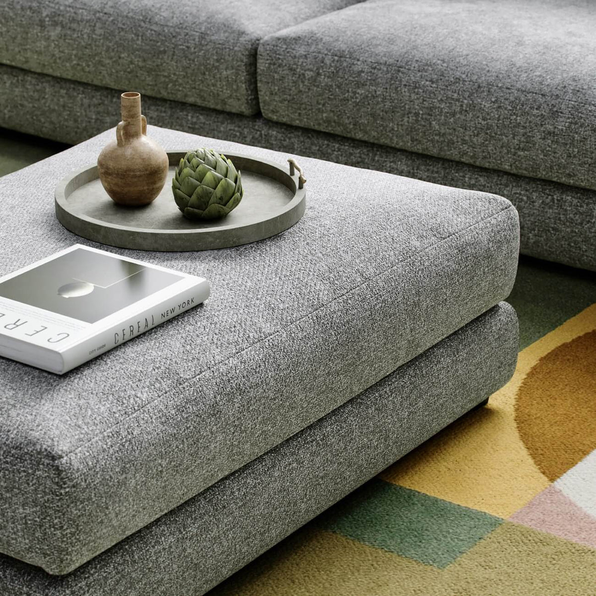 Modern gray ottoman with decorative tray, artichoke, ceramic vase and coffee table book on geometric rug
