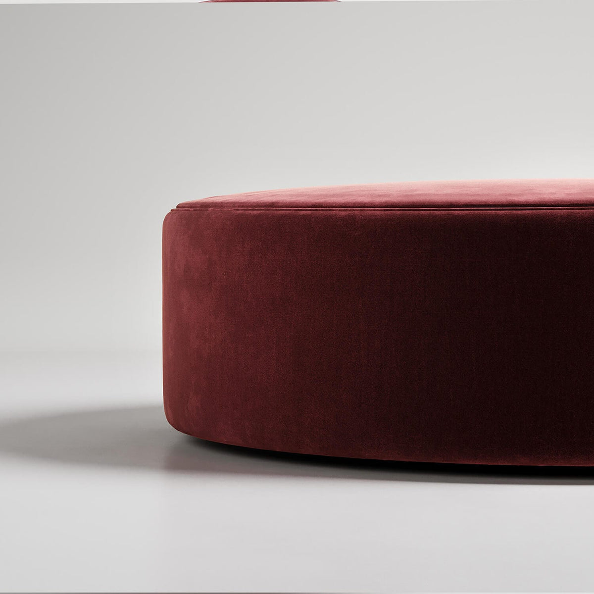Minimalist photo of round burgundy velvet ottoman against white background, showing curved edge detail
