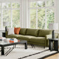 Modern living room with olive green sofa, black accent tables, geometric rug, white windows overlooking greenery
