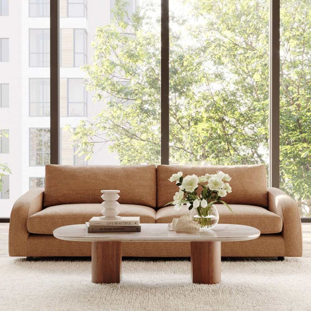 Modern brown sofa with oval coffee table, white flowers in vase, books near bright window with leafy view

